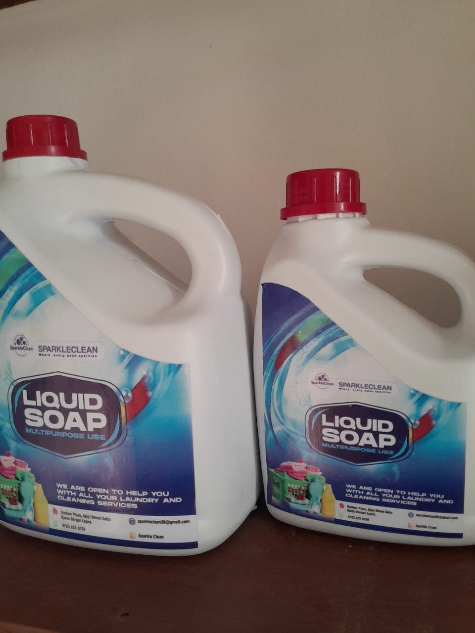 Two white plastic jugs of SparkleClean multipurpose liquid soap with red caps side-by-side.